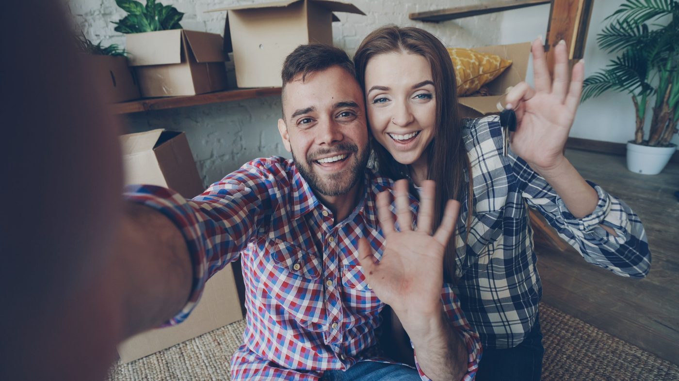 A smiling couple holding up their new house keys surrounded by moving boxes